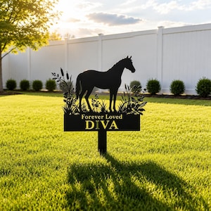 May include: A black metal garden stake featuring a silhouette of a horse standing among tall grasses and cattails. Below the horse, a rectangular plaque displays the text Forever Loved and the name DIVA in bold, capitalised yellow lettering.
