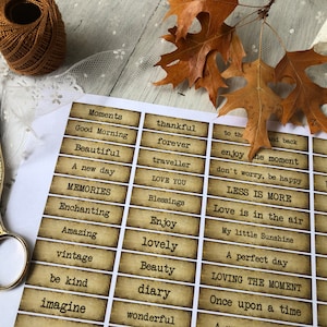 May include: A sheet of paper with vintage-style labels featuring inspirational words like "Moments," "Beautiful," and "Enjoy." The labels have a distressed, aged appearance. Gold scissors, brown thread, and autumn leaves are also present.