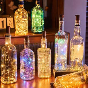 Puede incluir: Una colección de botellas de vidrio transparente, cada una con un tapón de corcho y rellena de luces de alambre. Las luces emiten varios colores, incluyendo amarillo cálido, azul frío, morado y verde, creando un efecto brillante.