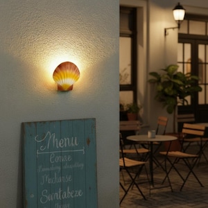 Op de afbeelding: Een wandlamp in de vorm van een schelp met een warme, gloeiende geel-oranje gradiëntafwerking. Onder de lamp staat op een verweerd blauwgroen houten bord het woord Menu in witte letters, gevolgd door verschillende regels tekst.