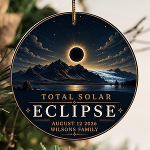 May include: A round ceramic ornament featuring a total solar eclipse over a mountain range and lake. The text reads TOTAL SOLAR ECLIPSE, AUGUST 12 2026, and WILSONS FAMILY in gold lettering. It hangs from a gold string against a dark blue sky.