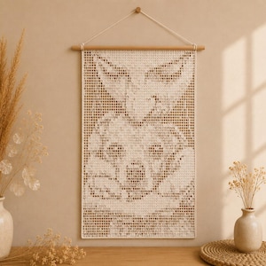 May include: A rectangular wall hanging featuring a grid-based crochet pattern of a rabbit face. The piece is suspended from a light-colored wooden dowel with a white cord. The design is created using off-white yarn, forming a pixelated image.