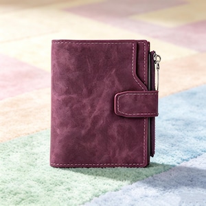 May include: A deep plum-colored bifold wallet with a textured matte finish. The wallet features a vertical zippered compartment on the right side, a snap-button closure strap, and visible tonal stitching along the edges of the exterior panels.