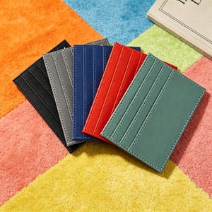 May include: A set of five rectangular card holders arranged in a fan shape. The holders are black, gray, blue, red, and sage green. Each holder features vertical stitched lines and multiple slots for organizing credit cards or identification.
