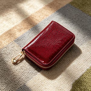 Puede incluir: Una cartera rectangular de color rojo oscuro con una textura brillante en relieve que imita la piel de cocodrilo. La cartera cuenta con una cremallera metálica de color dorado en tres lados y un tirador de metal dorado pulido.