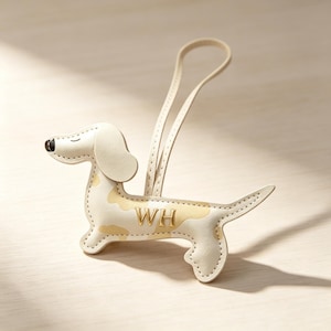 Puede incluir: Un colgante para bolso con forma de perro salchicha en color crema con manchas de color tostado claro, nariz negra y una correa. El costado del perro tiene grabadas las letras doradas WH. El accesorio presenta costuras visibles en los bordes.