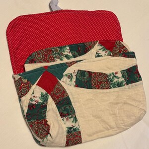 May include: A quilted fabric pouch featuring a red and white polka dot interior flap and a white elastic loop. The exterior displays a patchwork pattern of cream fabric, red paisley prints, and green holly leaf motifs with red berries.