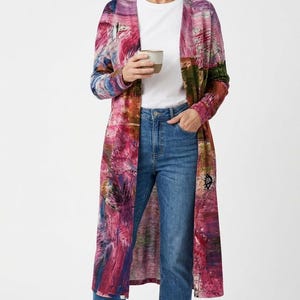 May include: A long, open-front cardigan featuring an abstract, painterly print in vibrant shades of magenta, pink, blue, and green. The garment is worn over a plain white crew-neck t-shirt and medium-wash blue denim jeans, paired with white sneakers.