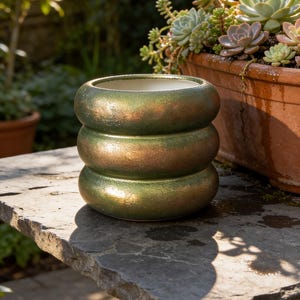 Pode incluir: Um vaso cilíndrico de cerâmica com três segmentos de anéis arredondados e empilhados com um acabamento metálico iridescente em tons de verde e bronze. O interior do vaso é liso e branco, contrastando com a superfície exterior texturizada.