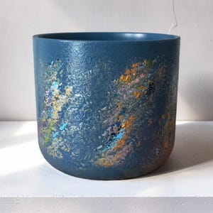 Può includere: Un vaso cilindrico con un colore di base blu profondo e una finitura testurizzata e maculata. L'esterno è decorato con schizzi di vernice multicolore sparsi in tonalità oro, argento, azzurro e rosa, creando un effetto a strati.