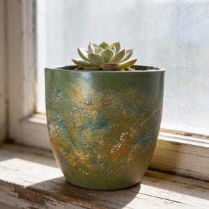 Peut inclure: Une petite plante succulente dans un pot en céramique vert olive. L'extérieur du pot présente une finition texturée et marbrée avec des touches mélangées de peinture dorée métallique, bleu sarcelle et blanche, créant un aspect abstrait.