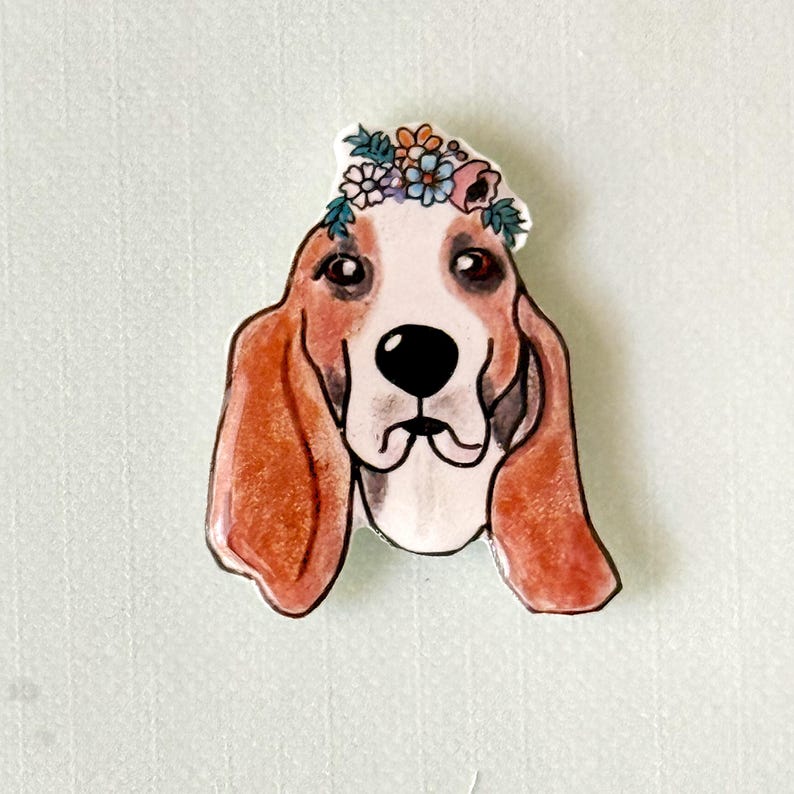 May include: A whimsical illustration of a Basset Hound wearing a floral crown. The dog has brown and white fur, with long, floppy ears. The floral crown features a variety of colorful flowers. The artwork is set against a light green background.