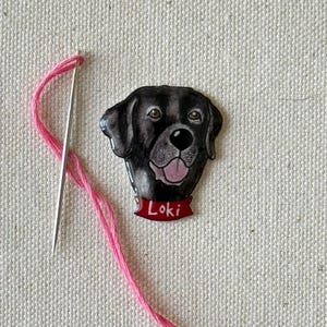 Puede incluir: Un parche bordado de la cabeza de un perro en blanco y negro, con el nombre "Loki" en una pancarta roja. El parche est&aacute; sobre un fondo de tela blanca con un hilo y una aguja rosas.