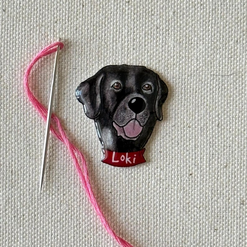 Puede incluir: Un parche bordado de la cabeza de un perro en blanco y negro, con el nombre "Loki" en una pancarta roja. El parche est&aacute; sobre un fondo de tela blanca con un hilo y una aguja rosas.