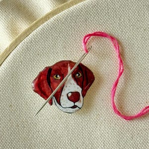 May include: A needle and pink thread are threaded through a brown and white dog head embroidery pattern. The dog has brown eyes and a white nose.