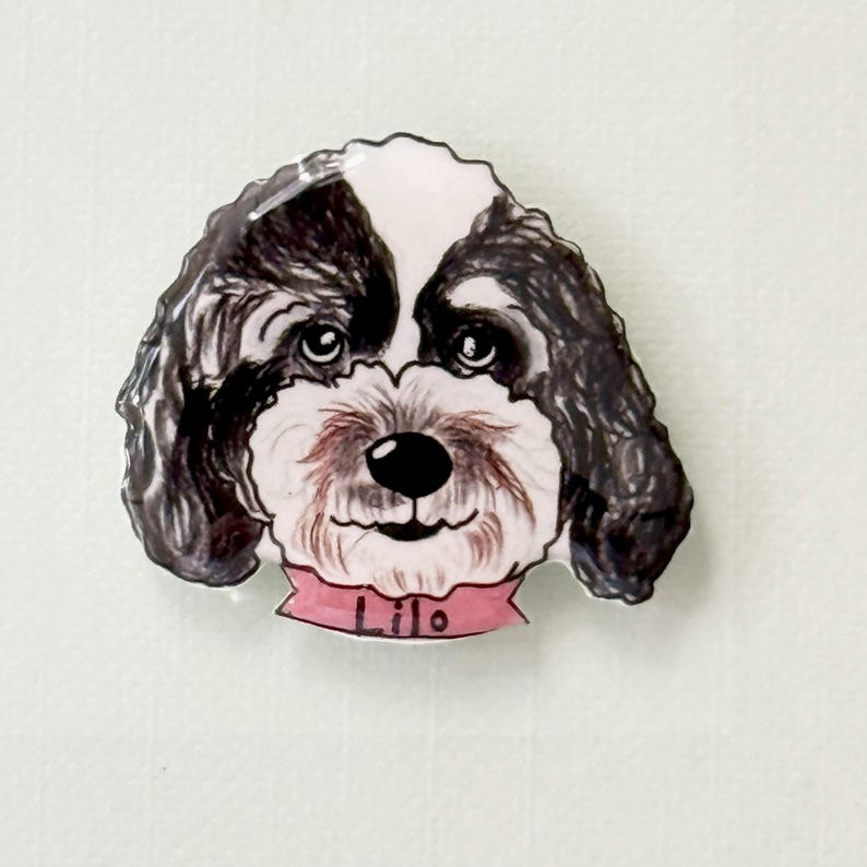 May include: A decorative magnet featuring a dog's face. The dog has black and white fur, a pink collar with the name "Lilo" printed on it, and a smiling expression. The magnet is likely for a refrigerator or other metal surface.