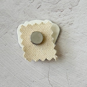 May include: A small, handmade magnet featuring a circular silver magnet adhered to a piece of cream-colored fabric with a textured, woven appearance. The fabric is attached to a white, irregular-shaped backing.