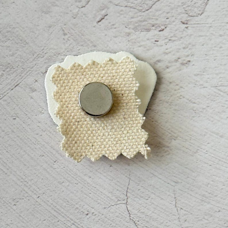 May include: A small, handmade magnet featuring a circular silver magnet adhered to a piece of cream-colored fabric with a textured, woven appearance. The fabric is attached to a white, irregular-shaped backing.