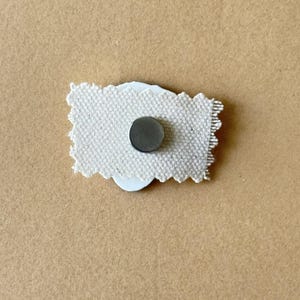 Puede incluir: Un parche de tela blanco con un im&aacute;n redondo plateado pegado en la parte posterior.