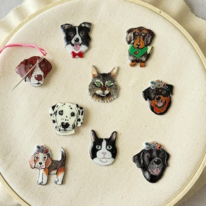 May include: A collection of pet-themed pins featuring various dog breeds and a cat. The pins are colorful and detailed, with each animal's unique features highlighted. The pins are arranged on a cream-colored fabric stretched within an embroidery hoop.