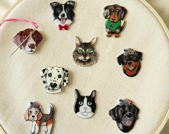 Puede incluir: Colección de coloridos pines con temática de mascotas, incluyendo perros y gatos, dispuestos sobre una tela crema dentro de un bastidor de bordado. Los pines presentan ilustraciones detalladas de varias razas, algunos adornados con accesorios.