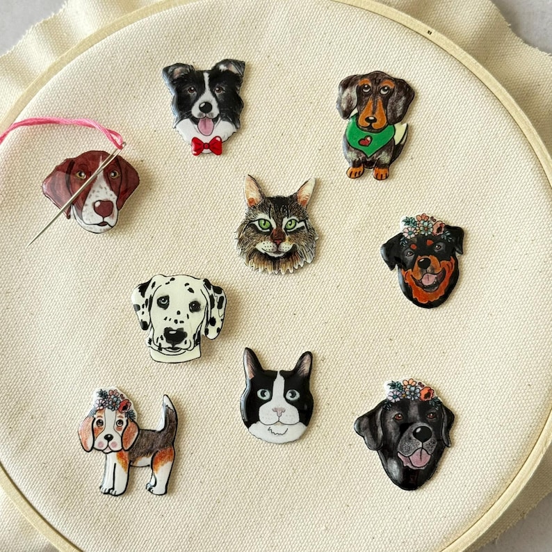 May include: A collection of pet-themed pins featuring various dog breeds and a cat. The pins are colorful and detailed, with each animal's unique features highlighted. The pins are arranged on a cream-colored fabric stretched within an embroidery hoop.