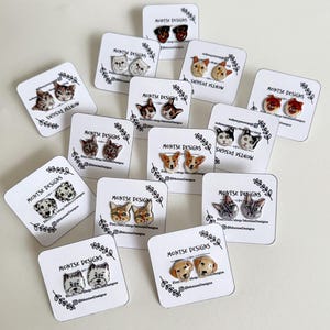 May include: A collection of pet-themed stud earrings, each featuring a different breed of dog or cat. The earrings are displayed on small white cards with the text "MONTSE DESIGNS". The earrings are in various colors and styles.