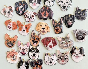 Puede incluir: Una colección de colgantes pintados a mano con caras de perros y gatos, con anillas doradas. Los colgantes presentan varias razas, como un dálmata, un husky y un carlino. La obra de arte es detallada, con pelaje y expresiones faciales realistas.