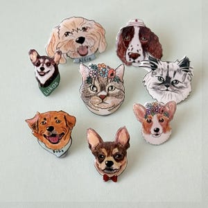 May include: A collection of colorful pet portrait pins featuring various dog and cat breeds. Each pin showcases a detailed illustration of a pet's face, with some adorned with floral crowns or accessories. The pins are set against a light green background.