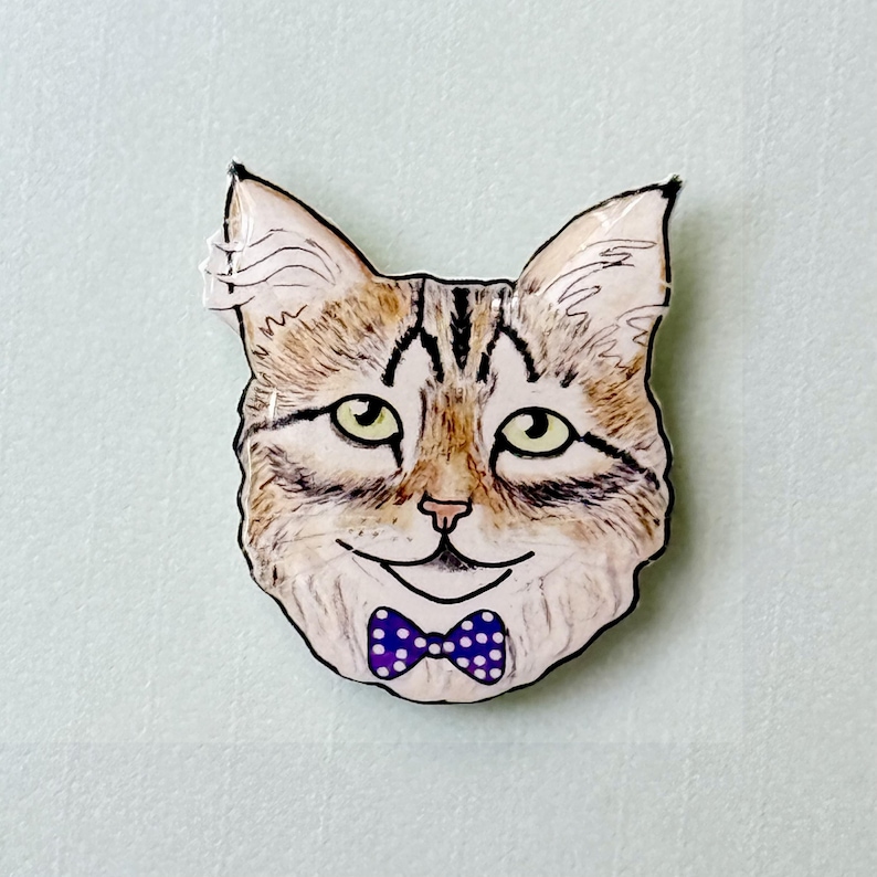 May include: A cat-shaped pin with a smiling face and a purple polka-dot bow tie. The cat has tan and brown fur, green eyes, and black outlines. The pin is set against a light blue background.