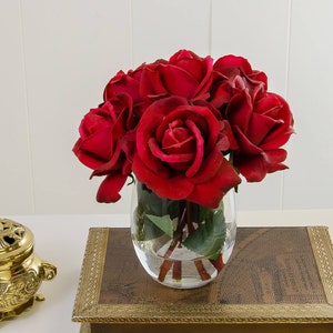 May include: A bouquet of red roses in a clear glass vase sits on a brown wooden box with gold trim. The roses are arranged in a circular shape and are in full bloom.