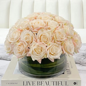 May include: A bouquet of blush pink roses in a clear glass vase with green leaves. The vase is sitting on a white marble coffee table with a book titled "Live Beautiful" by Caldeone.