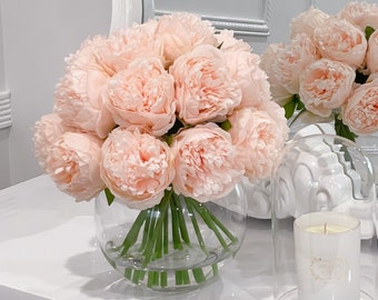 May include: A white vanity table with a large ornate mirror. A bouquet of pink peonies in a clear glass vase sits on the table, along with a white candle in a clear glass cloche.