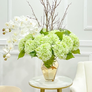 Könnte beinhalten: Ein weißer und grüner Blumenstrauß in einer goldenen geometrischen Vase. Die Vase steht auf einem weißen Marmortisch mit goldenen Beinen. Der Strauß enthält weiße Orchideen und grüne Hortensien.