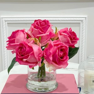 May include: A bouquet of pink roses in a clear glass vase. The roses are arranged in a cluster with their stems visible. The vase is sitting on a pink surface.