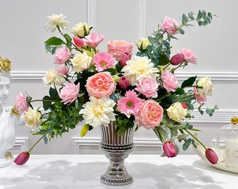 May include: A silver vase filled with a large arrangement of pink and white flowers. The flowers include roses, gerberas, and dahlias. The vase is on a white table.