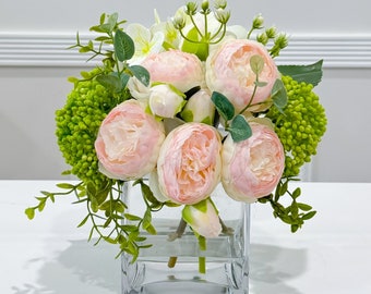 May include: A bouquet of pink and white artificial flowers in a clear glass vase. The flowers are arranged in a cluster and include peonies and greenery.