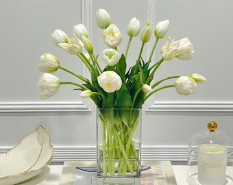May include: A bouquet of white tulips in a clear glass vase on a table with books. The books have the titles "Live Beautiful" and "White".