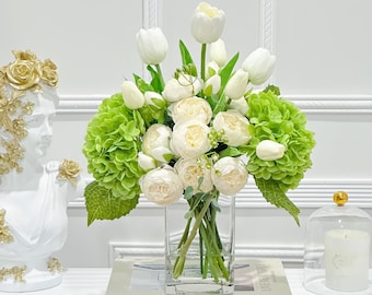 May include: A white vase filled with white tulips and white ranunculus flowers, accented with green hydrangea blooms. The vase is sitting on a white surface with a book titled "Live Beautiful White" by Chrissie Rucker.