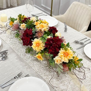 Puede incluir: Una mesa de comedor con un centro de mesa floral. El arreglo presenta hortensias burdeos, dalias amarillas y rosas melocotón. Se colocan platos blancos, cubiertos y copas de vino. Un camino de mesa beige está en el centro.