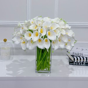 May include: A display of white calla lilies with yellow centres and green stems, presented in a clear, rectangular glass vase. The flowers are arranged on a white surface, with a glass cloche and books in the background.