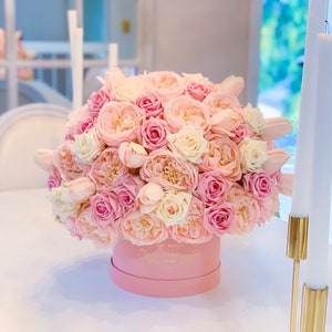 Puede incluir: Una caja rosa llena de un gran ramo de rosas rosas y blancas. La caja tiene un logotipo dorado que dice "The Roses Flowery".