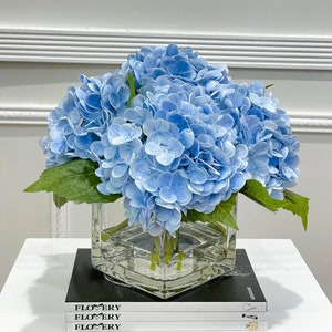May include: A bouquet of light blue hydrangeas in a clear glass cube vase. The vase is sitting on a stack of three books with the word "FLOWERY" printed on the covers.