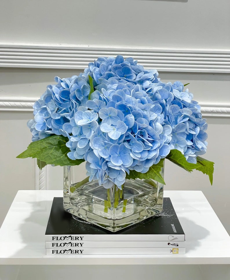 May include: A bouquet of light blue hydrangeas in a clear glass cube vase. The vase is sitting on a stack of three books with the word "FLOWERY" printed on the covers.