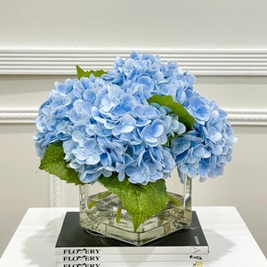 May include: A bouquet of light blue hydrangeas in a clear glass vase on a white table with a stack of black books that say "FLOWERY" on the cover.