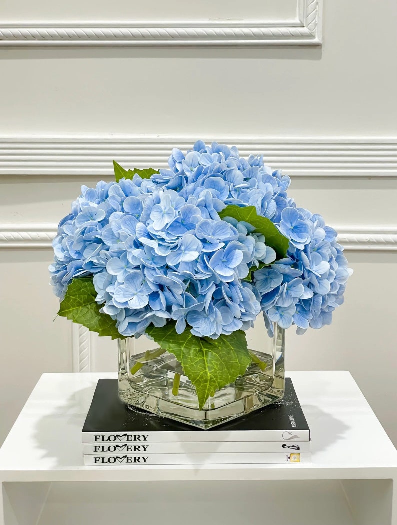 May include: A bouquet of light blue hydrangeas in a clear glass vase on a white table with a stack of black books that say "FLOWERY" on the cover.