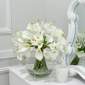 May include: A white vase filled with a bouquet of white calla lilies. The vase is sitting on a white table with a white candle in front of it.