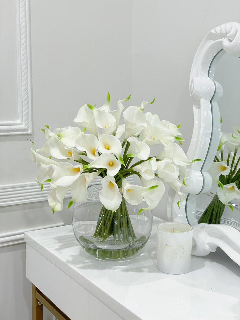 May include: A white vase filled with a bouquet of white calla lilies. The vase is sitting on a white table with a white candle in front of it.
