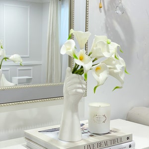 May include: White ceramic vase shaped like a hand holding a bouquet of white calla lilies. The vase is on a white table with two books stacked on top of each other. The top book has the title "Live Beautiful White" on the cover.