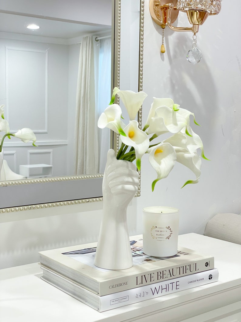 May include: White ceramic vase shaped like a hand holding a bouquet of white calla lilies. The vase is on a white table with two books stacked on top of each other. The top book has the title "Live Beautiful White" on the cover.
